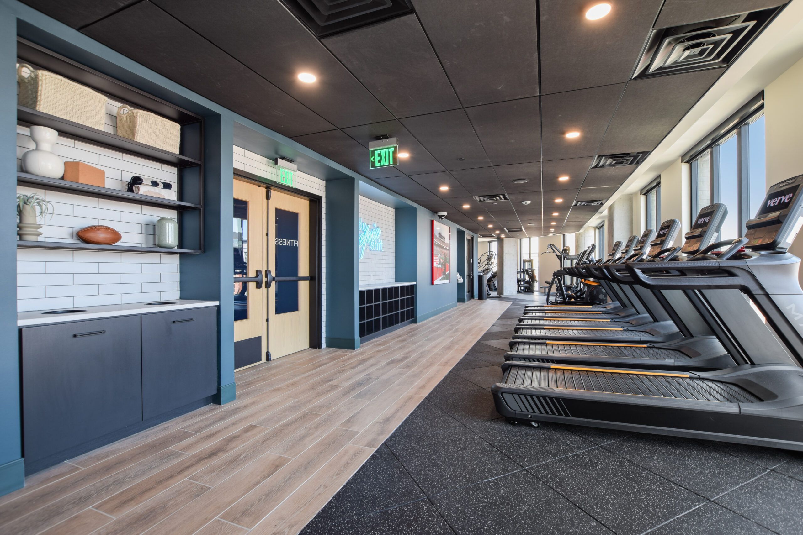 Modern gym with treadmills, wooden floor, ceiling lights, and decorated shelves.