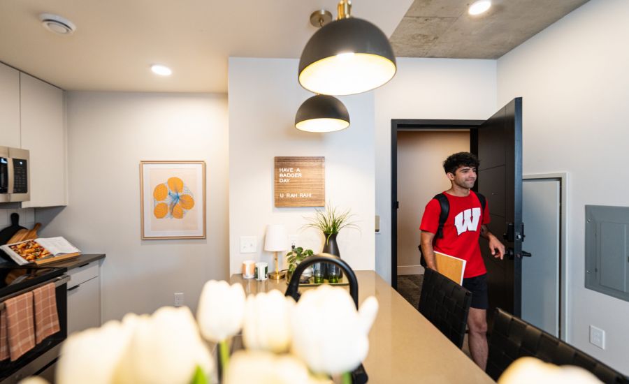 A man in a red shirt enters a modern kitchen with pendant lights and white tulips on the counter.