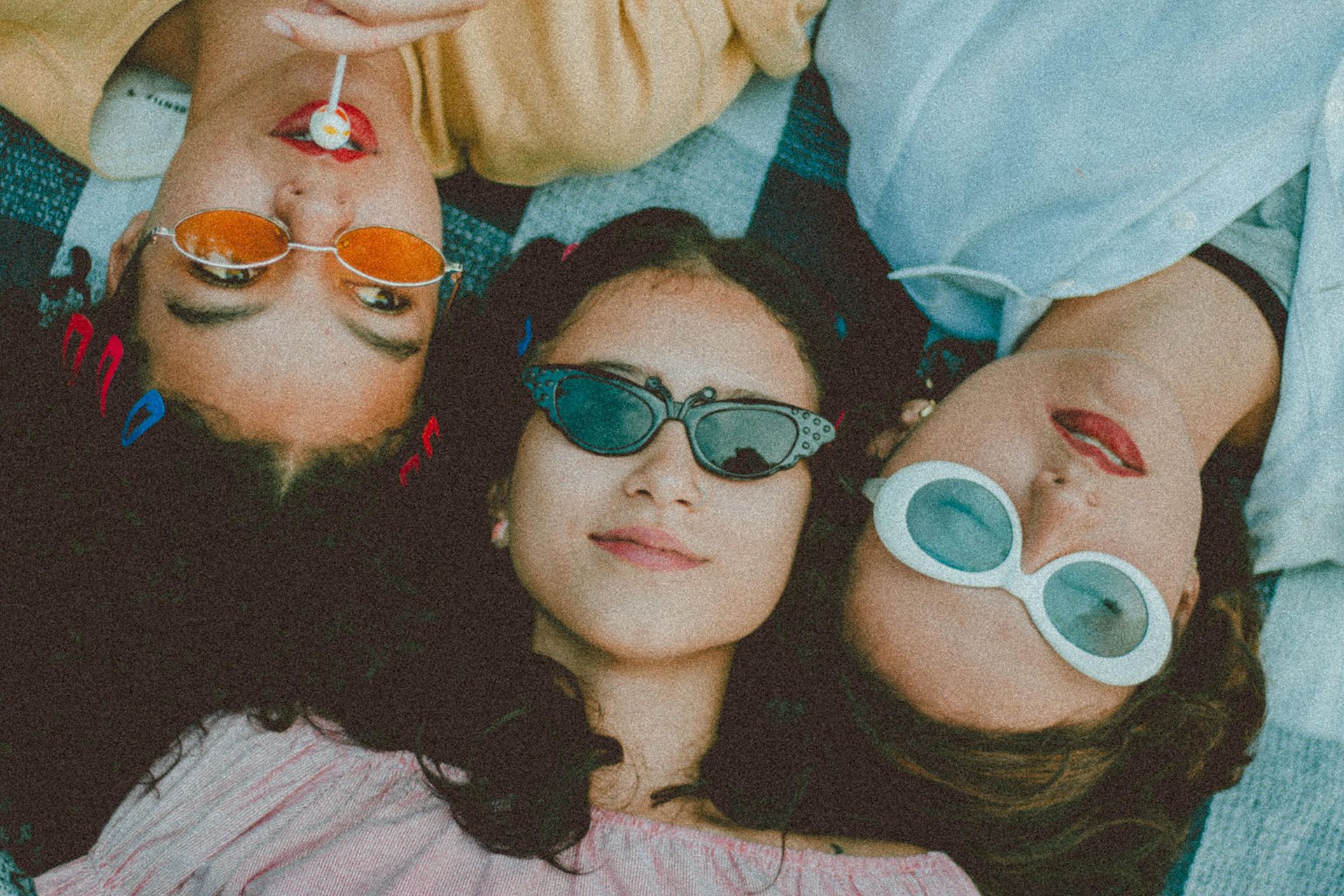 Three college aged girls laying on the ground with their heads alternating and wearing sunglassses
